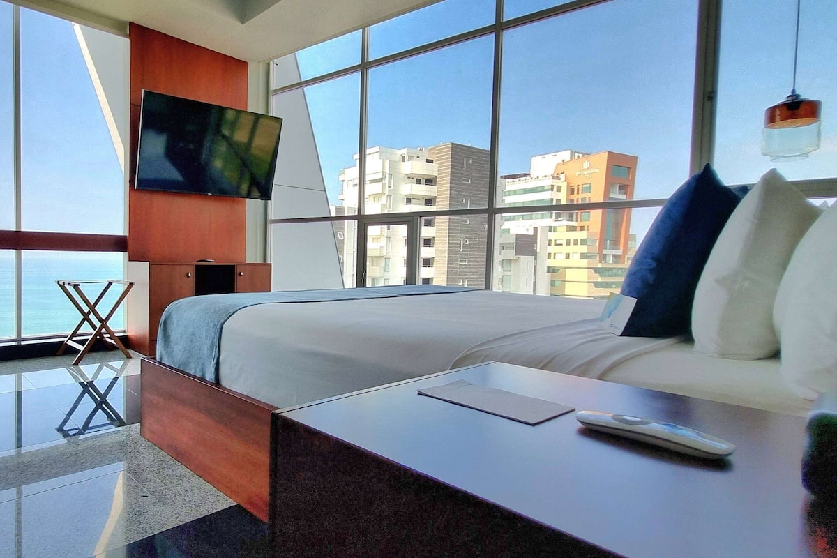 Une chambre d’hôtel moderne, dotée d’un grand lit, d’un téléviseur fixé au mur et offrant une vue sur le paysage urbain grâce à de grandes fenêtres.
