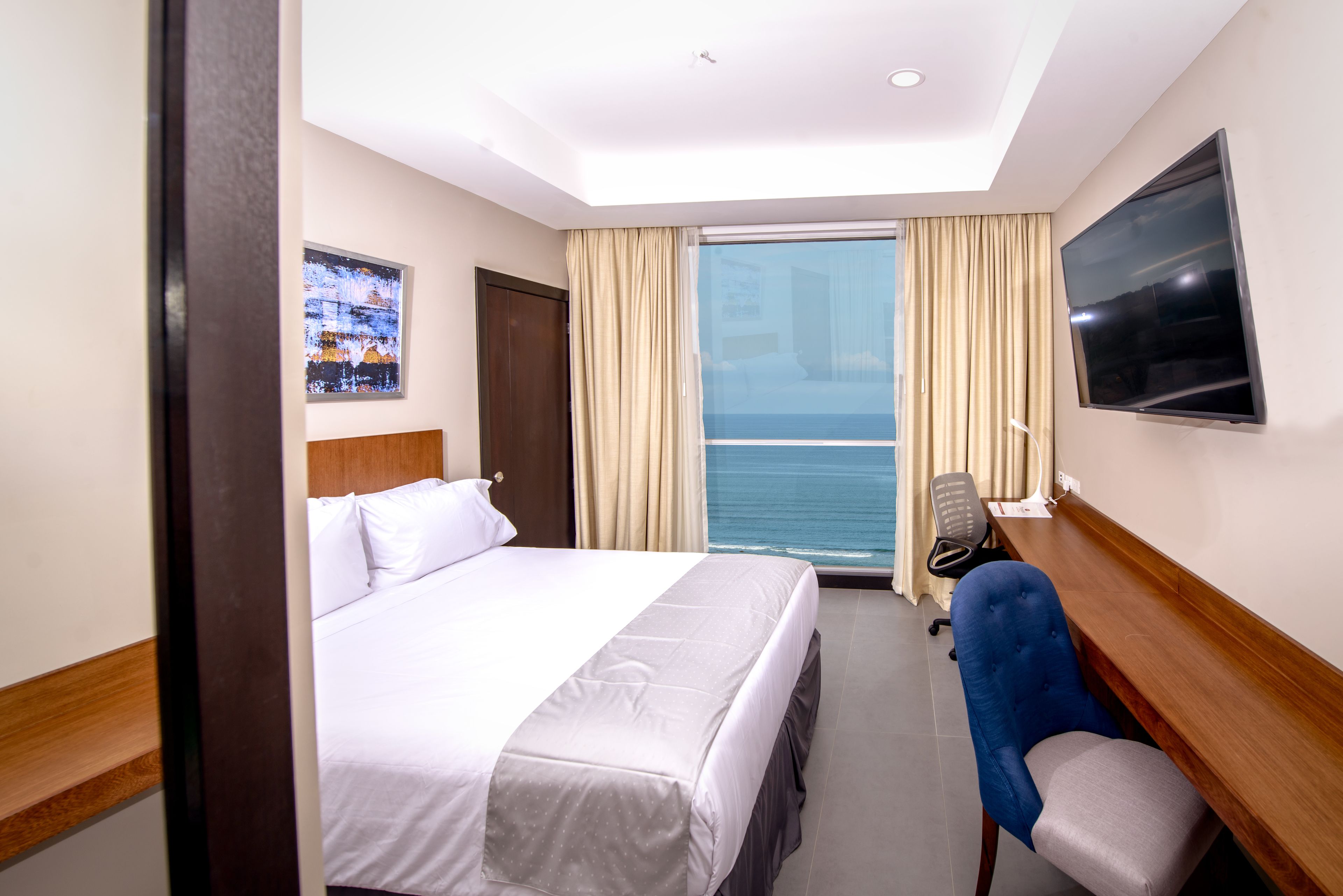 Habitación de hotel con cama, escritorio, silla, televisor y ventana con cortinas.