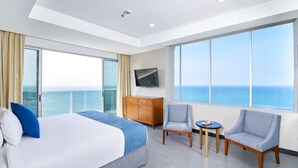 Una habitación de hotel moderna con una cama grande, televisión, una silla, una mesita y vistas al océano.