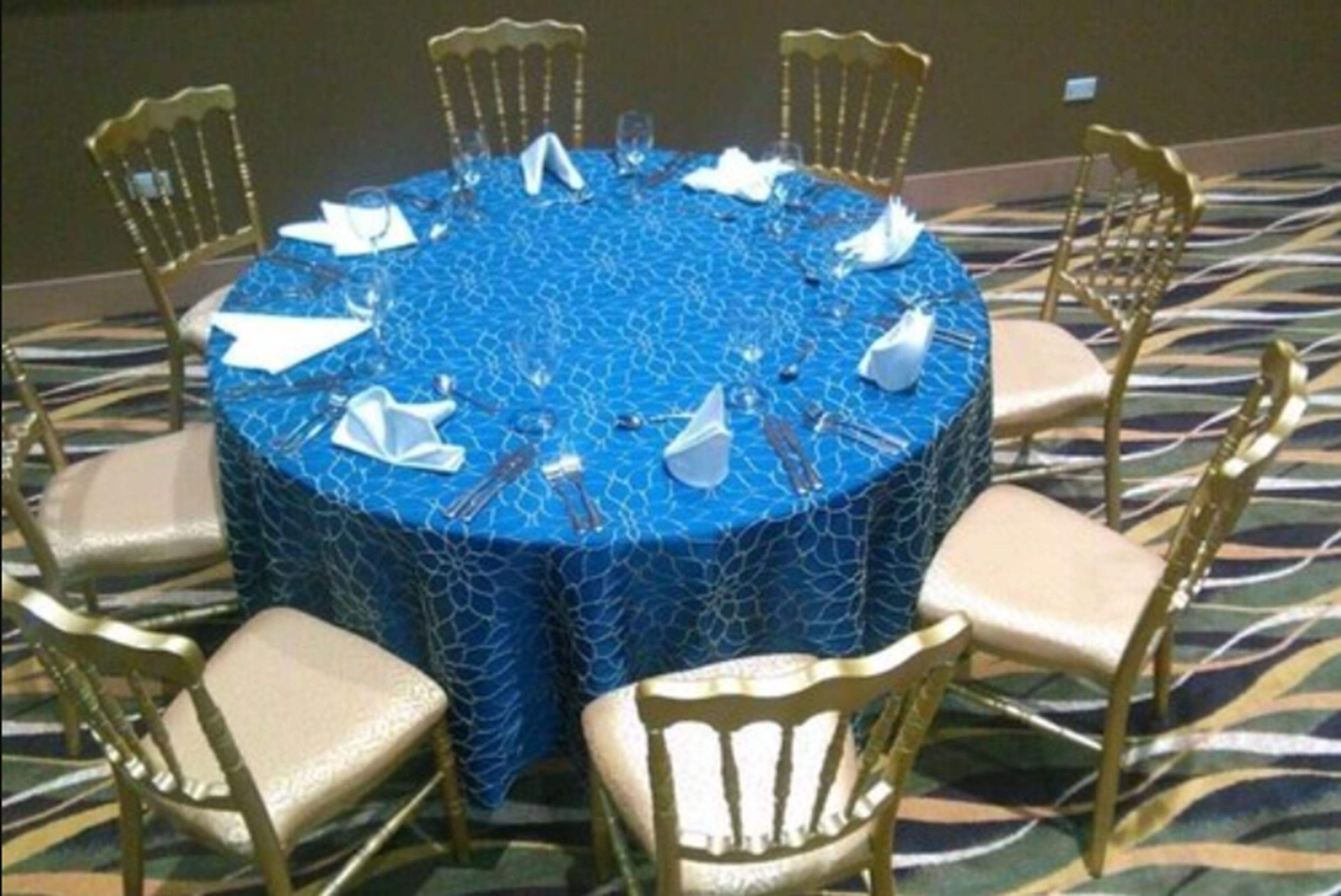 Una mesa redonda con mantel azul estampado, rodeada por seis sillas doradas con cojines color beige.