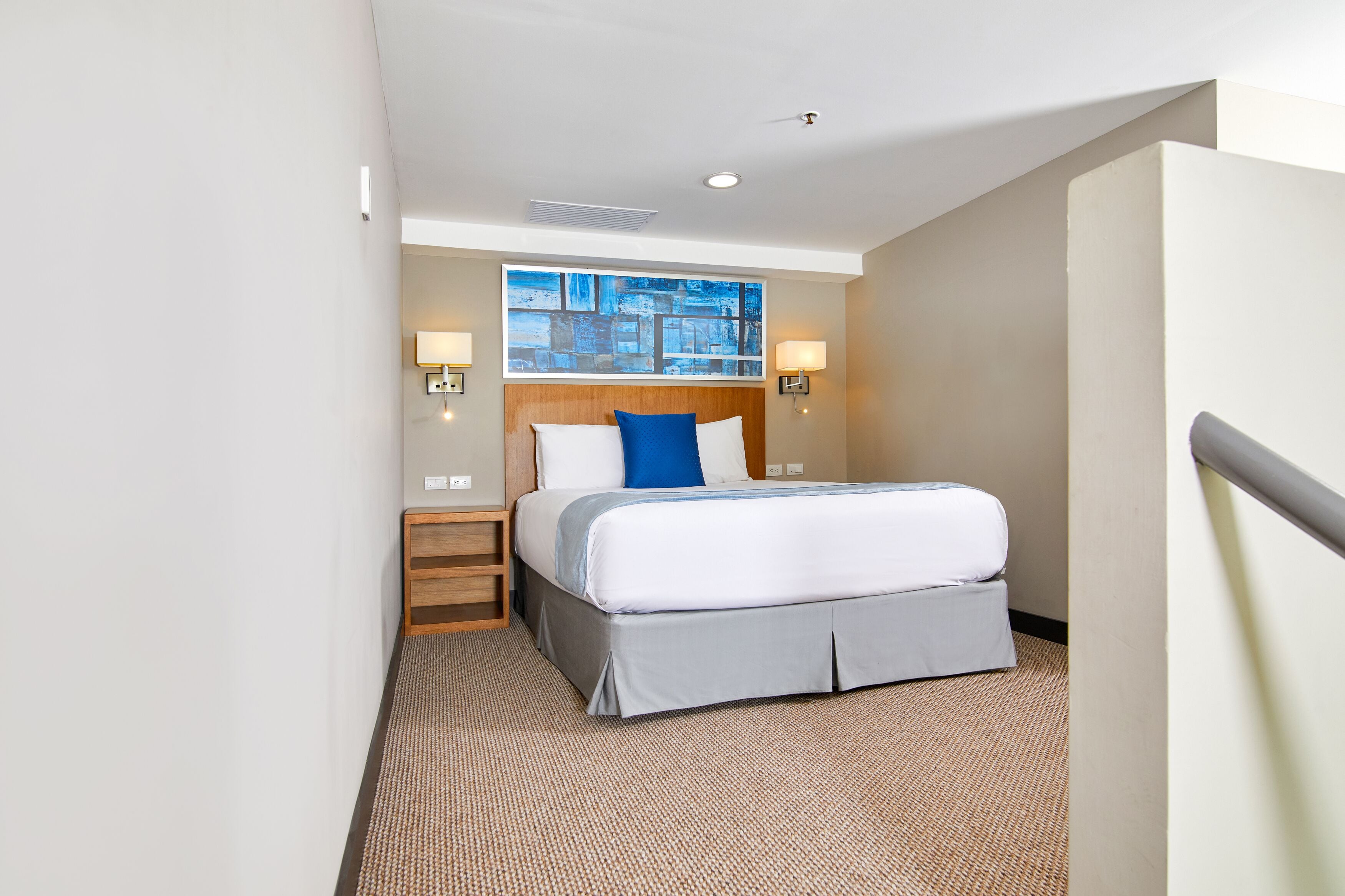 A hotel room with a bed, a painting on the wall, and two wall-mounted lamps.