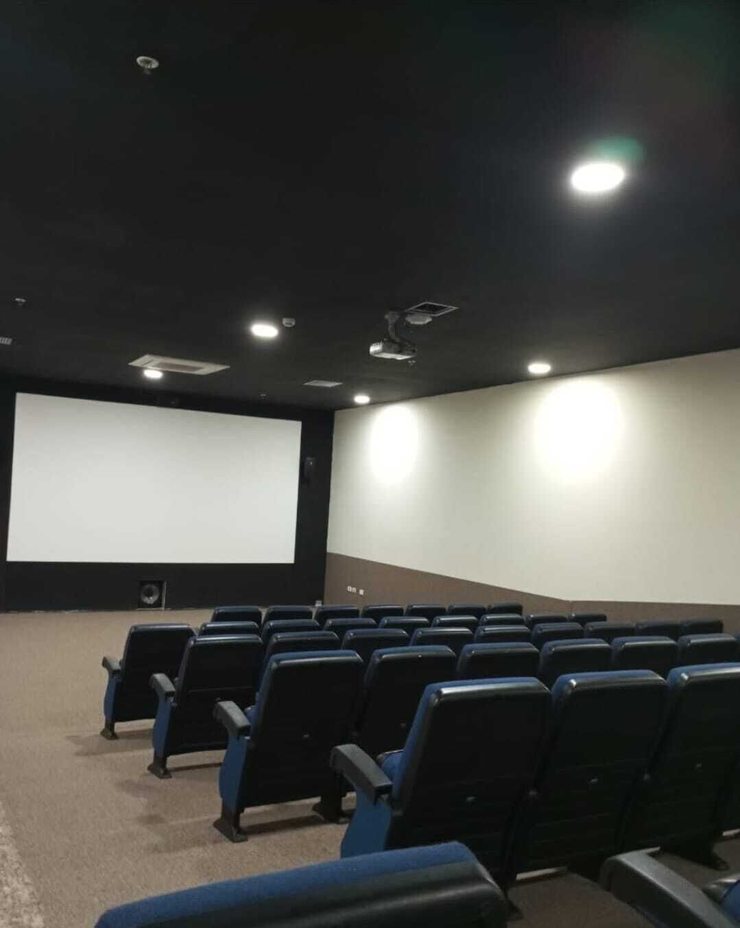 A movie theater with rows of black seats, a large screen, and ceiling lights.