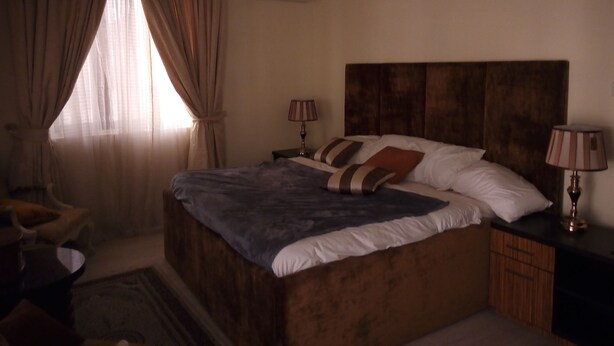 A bedroom with a large bed, two bedside tables with lamps, a chair, and a window with curtains. - Blossom Court (Ibadan)