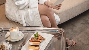 Une personne vêtue d’un peignoir blanc est assise sur un canapé beige et lit un livre. Un plateau avec une tasse de café et une assiette de nourriture se trouve devant elle.