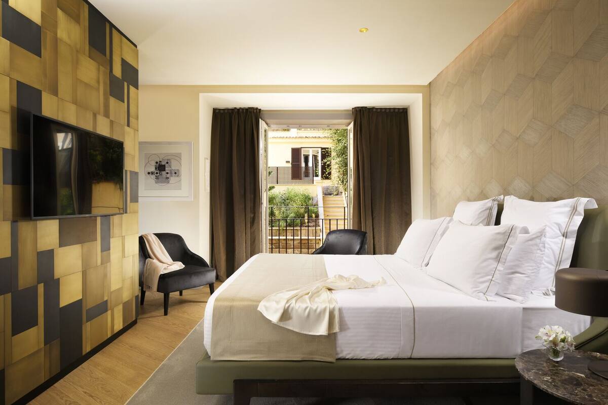 Una habitación de hotel moderna con una cama grande, un televisor montado en la pared, un área de descanso con una silla y vistas al balcón exterior.