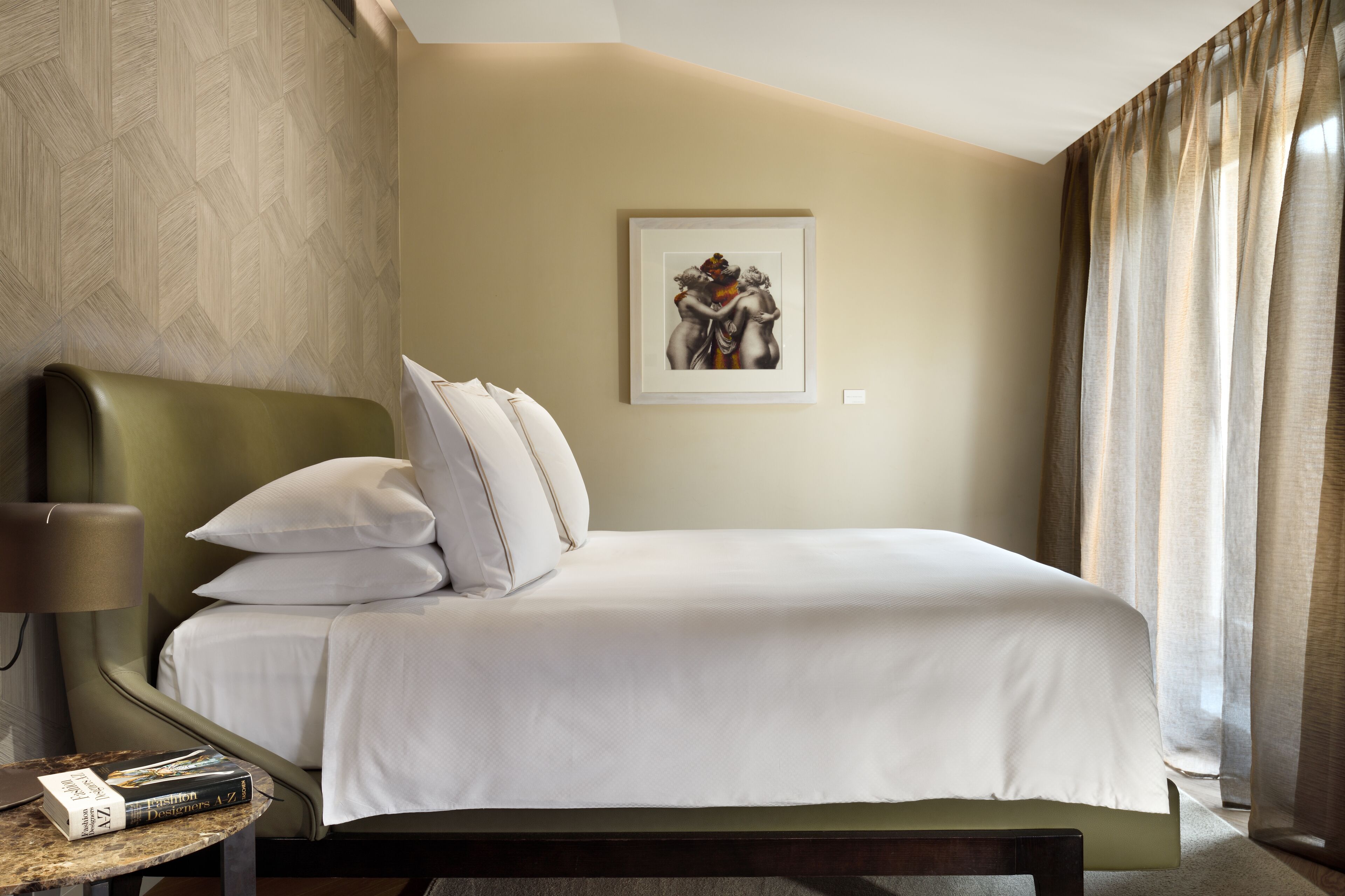 Une chambre d’hôtel avec un lit, des oreillers, une table de chevet avec une lampe, une œuvre d’art encadrée au mur et une fenêtre avec des rideaux.