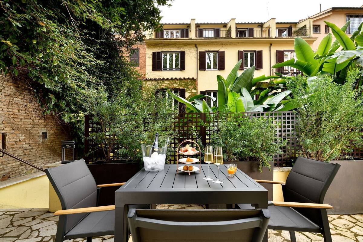 Un patio al aire libre con una mesa dispuesta para dos personas, rodeado de sillas. Hay una botella de champán, una copa de vino y un vaso de jugo de naranja sobre la mesa.