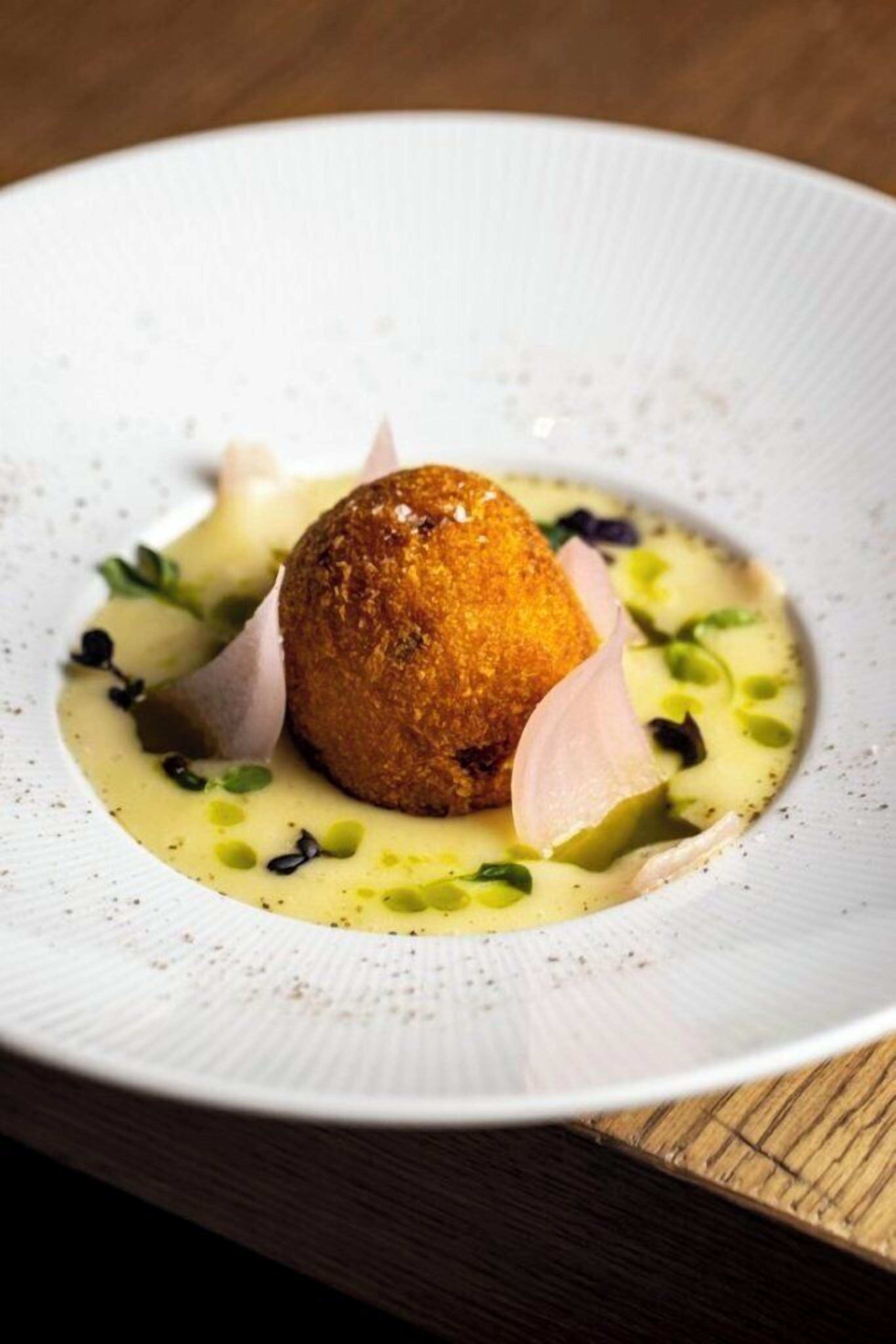 Ein weißer Teller mit einem einzelnen, rundlichen, goldbraunen Gericht, möglicherweise frittiert, umgeben von grüner Soße und dünn geschnittenen Zwiebeln.