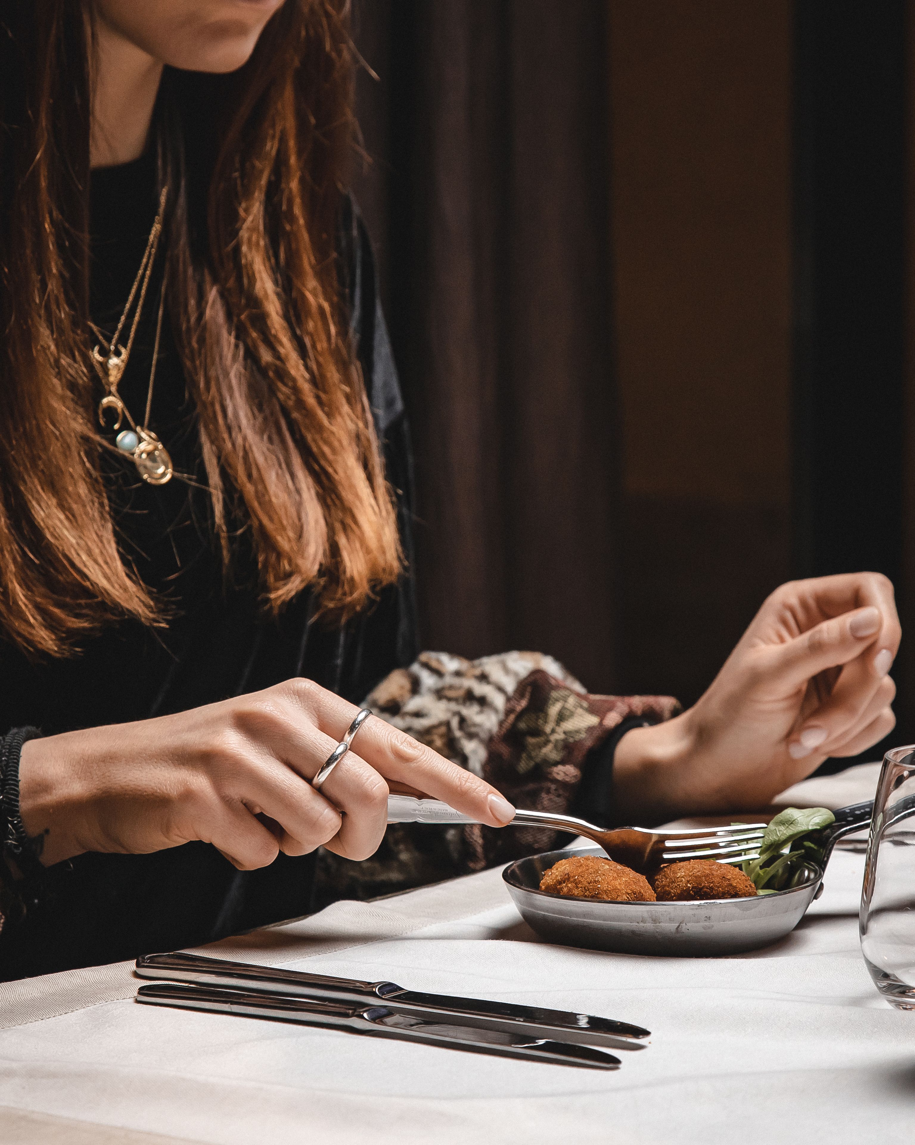 Une personne est assise à une table, devant une assiette de nourriture, des couverts et un verre d’eau.