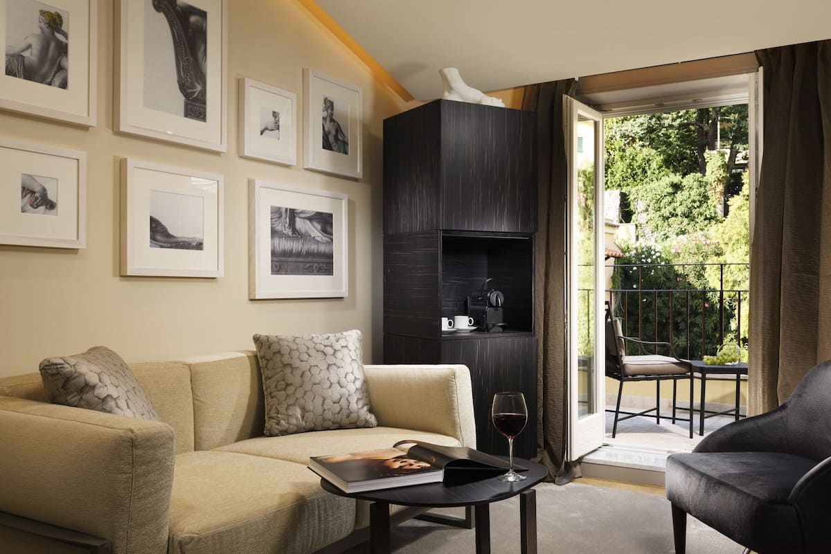 Una sala de estar con un sofá, una mesa baja con un libro y una copa de vino, un mueble de madera oscura y un balcón con una silla y una mesa.