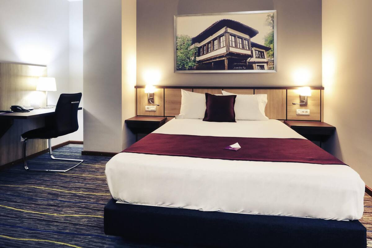 A hotel room with a bed, a desk with a chair, two bedside lamps, and a painting on the wall.