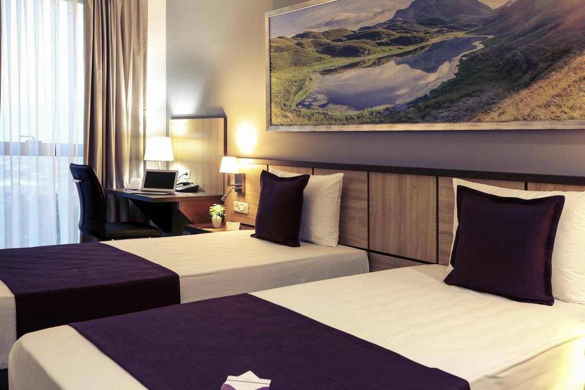 A hotel room with two beds, a desk with a laptop, a lamp, and a painting of a landscape.
