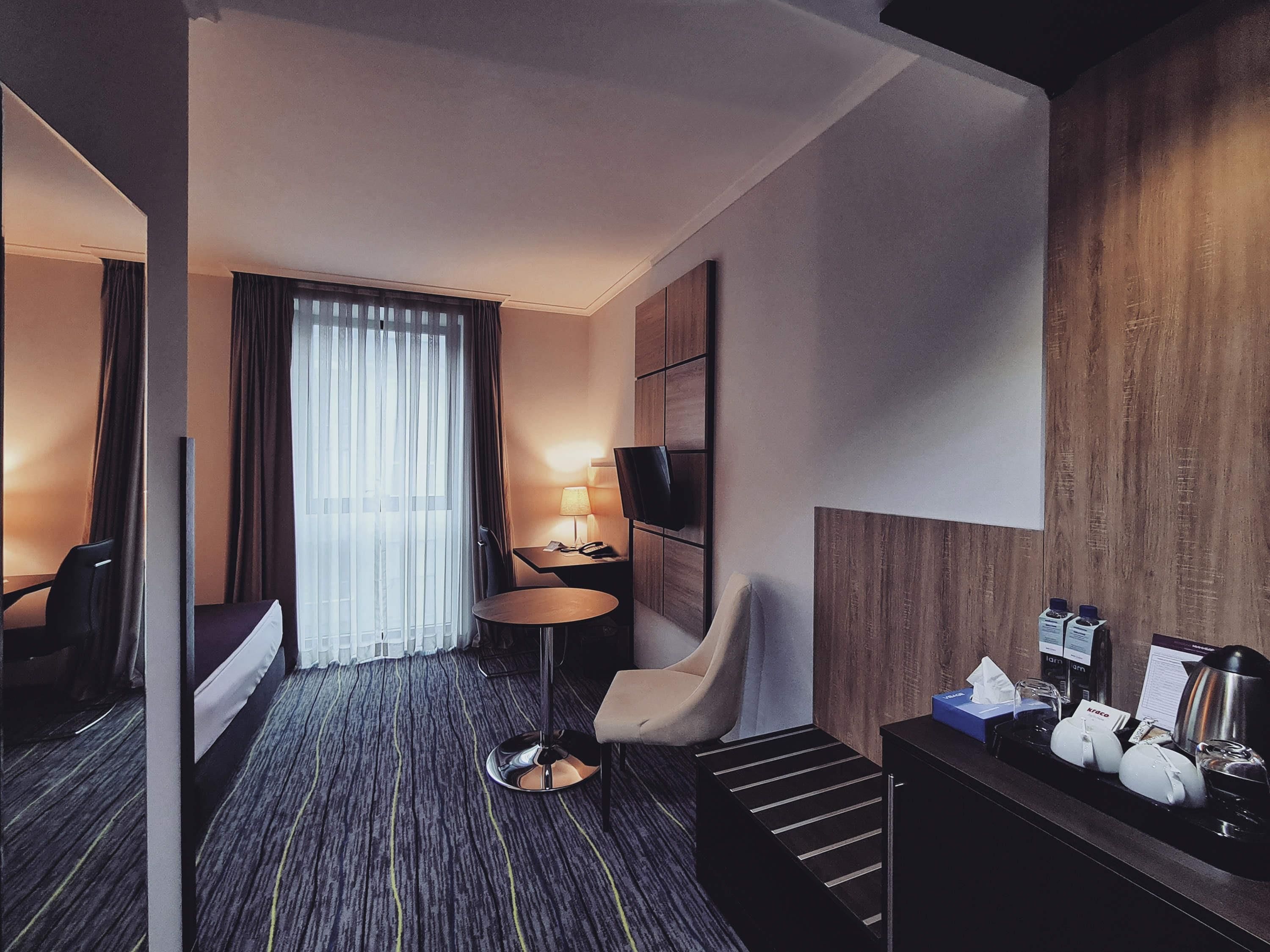 A hotel room with a bed, a chair, a small table with a lamp, a TV mounted on the wall, and a minibar.