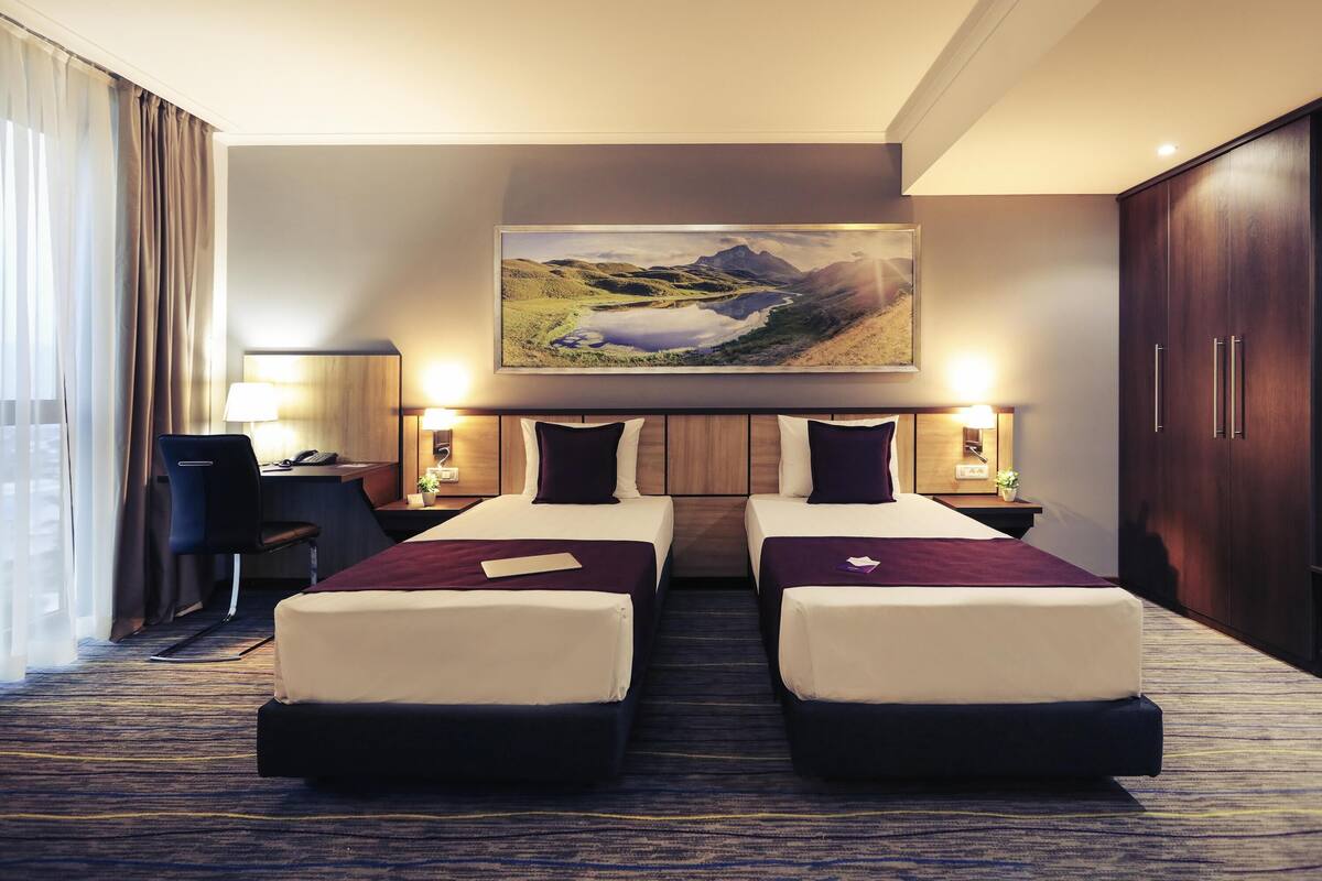 A hotel room with two beds, a desk, a chair, a lamp, and a painting of a landscape.