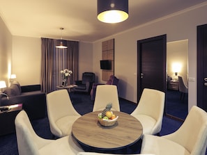 A hotel room with a round table, two armchairs, a sofa, a TV, a lamp, and a door.