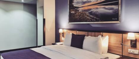Una habitación de hotel con una cama grande, una mesita de noche con una lámpara, un cuadro colgado en la pared y una puerta hacia otra habitación.