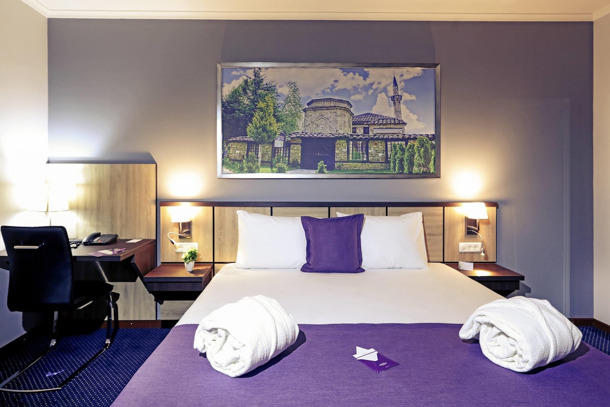Une chambre d’hôtel avec un lit, deux tables de chevet, un bureau et une chaise. Il y a deux serviettes sur le lit et un tableau au-dessus du lit.