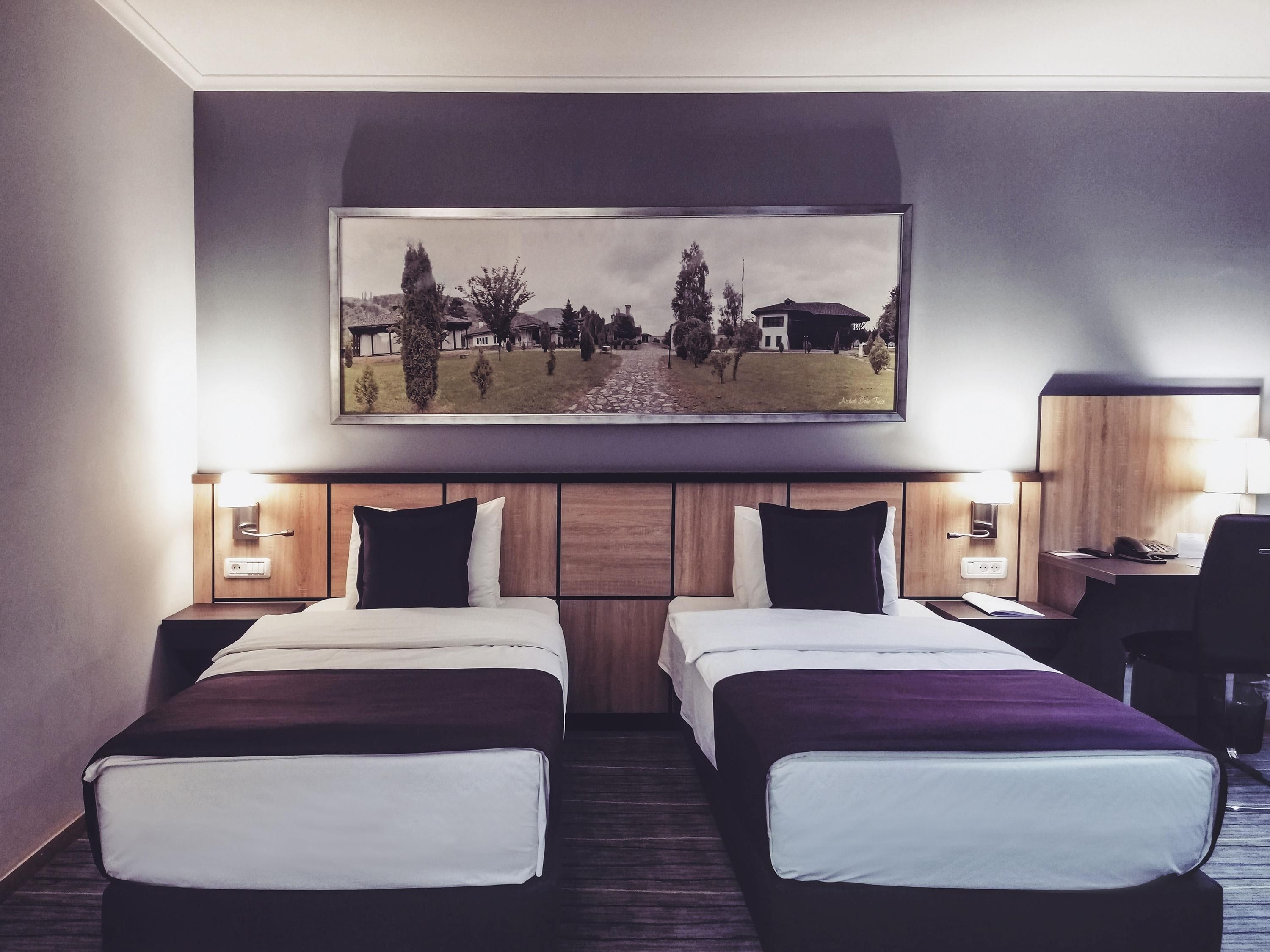 A hotel room with two beds, a desk, a chair, a painting above the beds, and two bedside lamps.