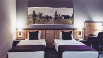 A hotel room with two beds, a desk, a chair, a painting above the beds, and two bedside lamps.
