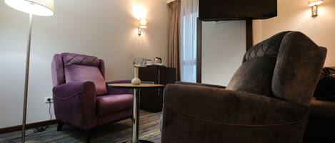 Quarto de hotel com uma poltrona roxa, uma poltrona marrom, uma mesinha, um candeeiro de chão e uma televisão de tela plana fixada na parede.
