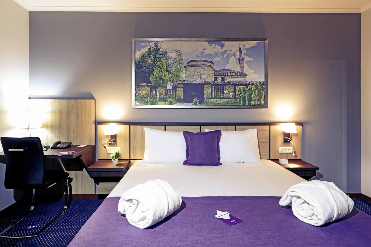 A hotel room with a bed, two nightstands, a desk, and a chair. There are two towels on the bed and a painting above the bed.