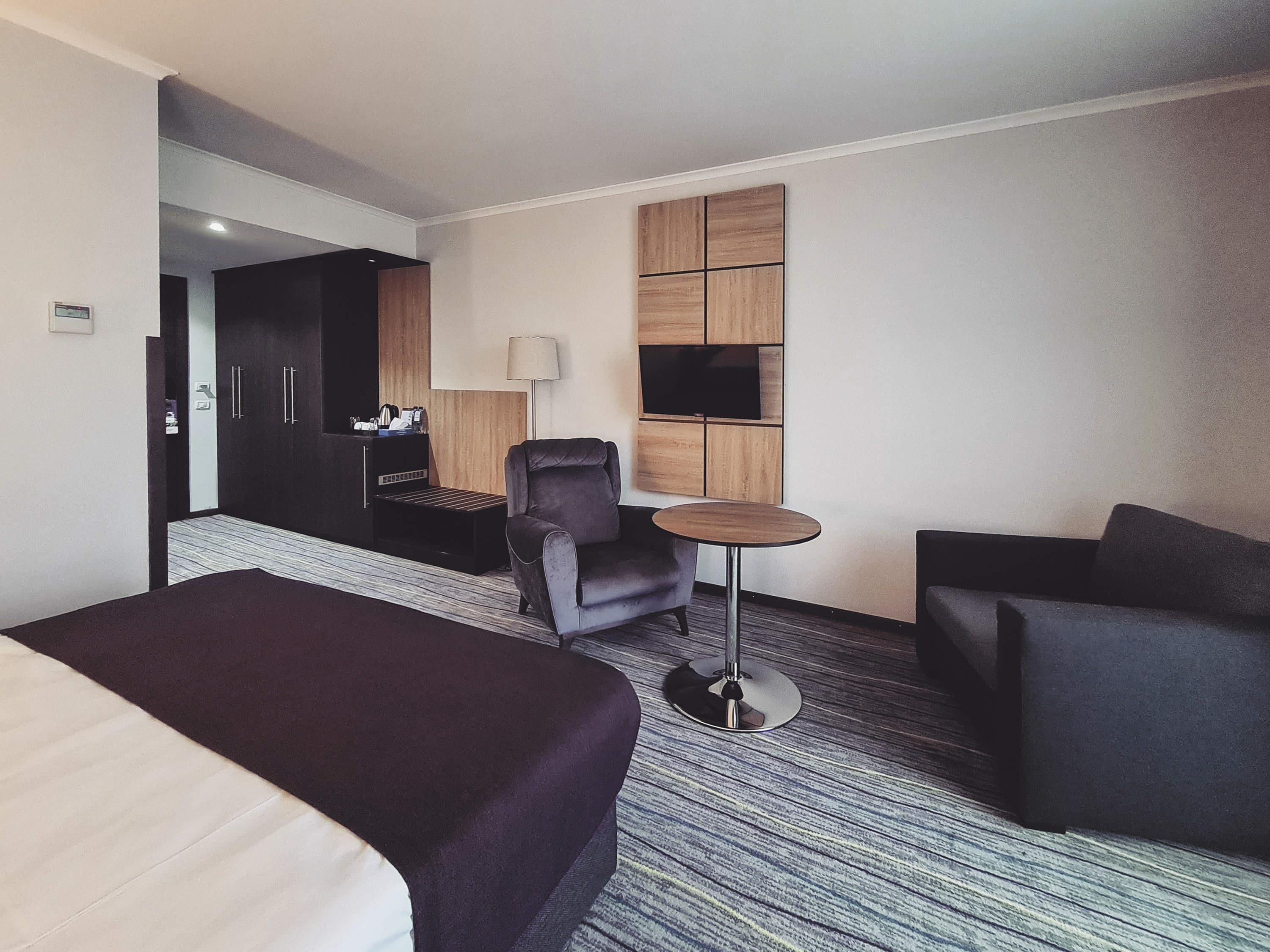 A hotel room with a bed, two armchairs, a small round table, a TV mounted on a wooden panel wall, and a wardrobe with a mirror.