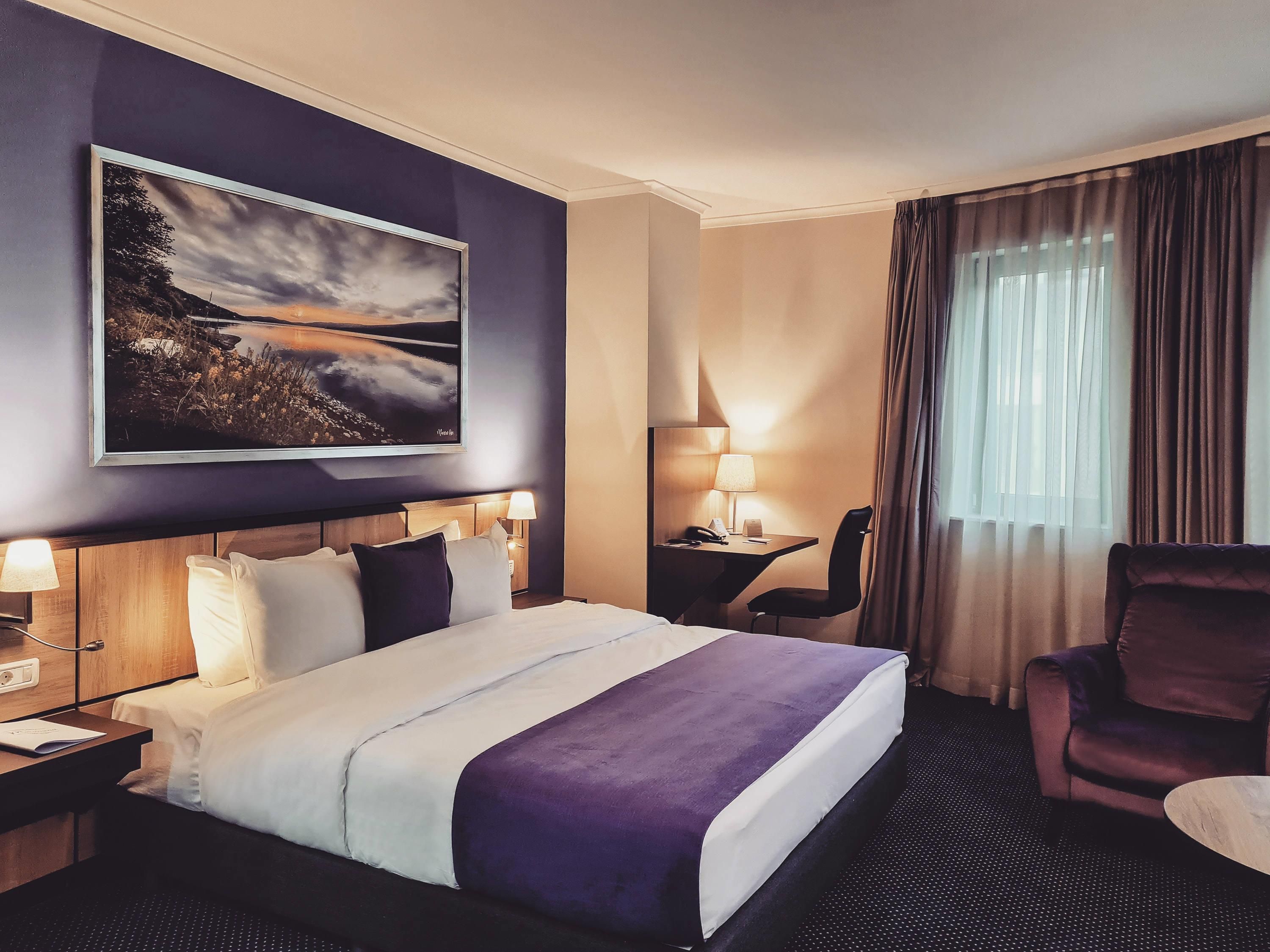 Una habitación de hotel con cama, escritorio, silla, lámpara y un cuadro en la pared.
