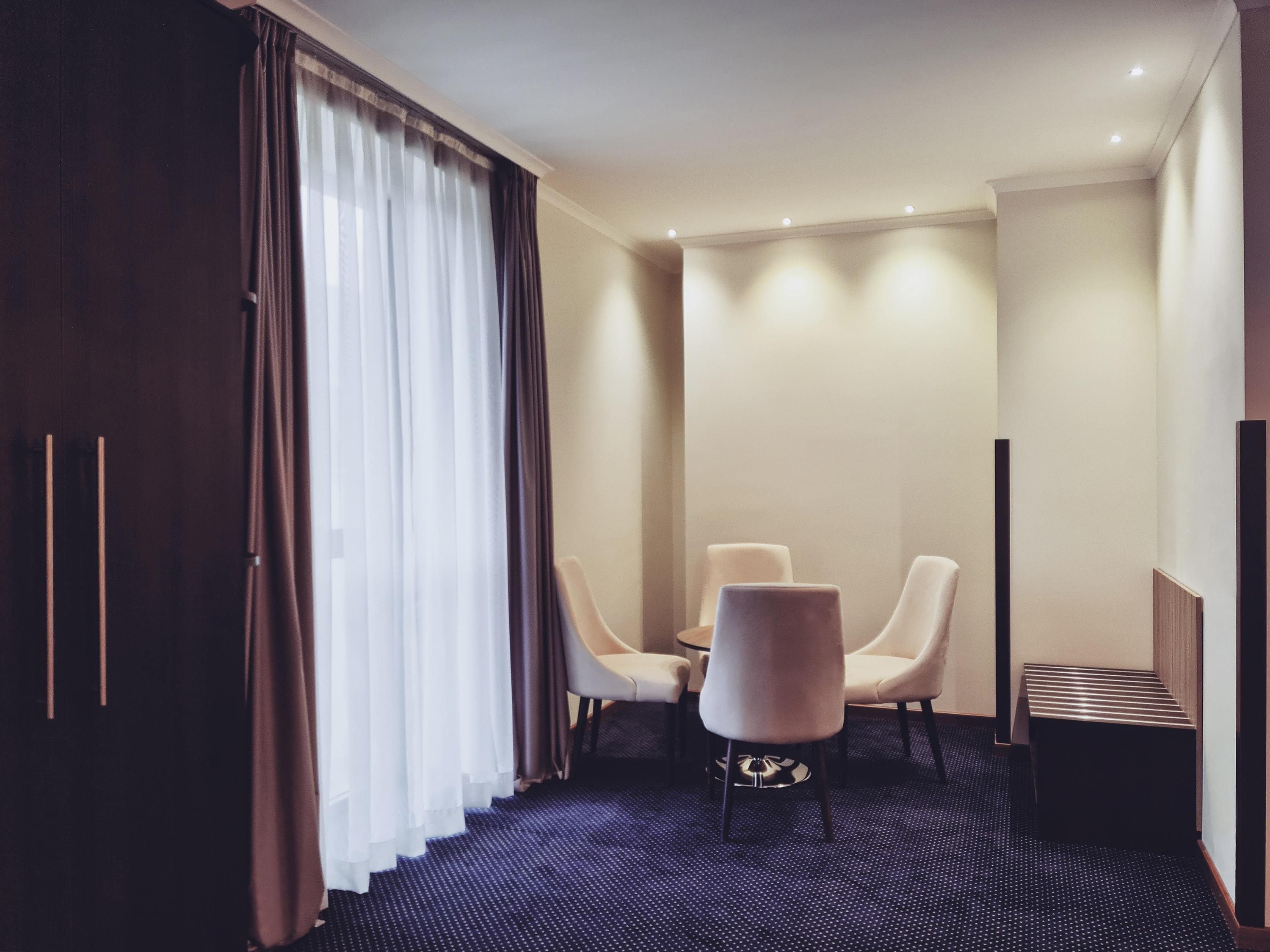 A hotel room with a dark carpet, a dining table with four chairs, a TV stand, and a large window with curtains.