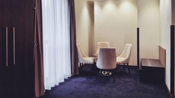 A hotel room with a dark carpet, a dining table with four chairs, a TV stand, and a large window with curtains.