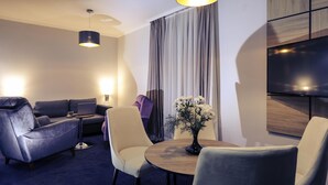 Une chambre d’hôtel moderne équipée d’un canapé, de fauteuils, d’une table à manger, d’un téléviseur et d’une grande fenêtre avec des rideaux.