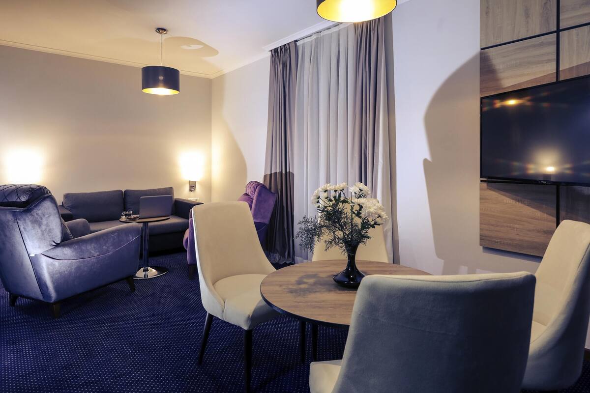 A modern hotel room with a sofa, chairs, a dining table, a TV, and a large window with curtains.
