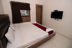 Uma cama de solteiro com estrutura de madeira, um tapete vermelho e roupa de cama branca. Há uma TV fixada na parede e uma janela com cortina.
