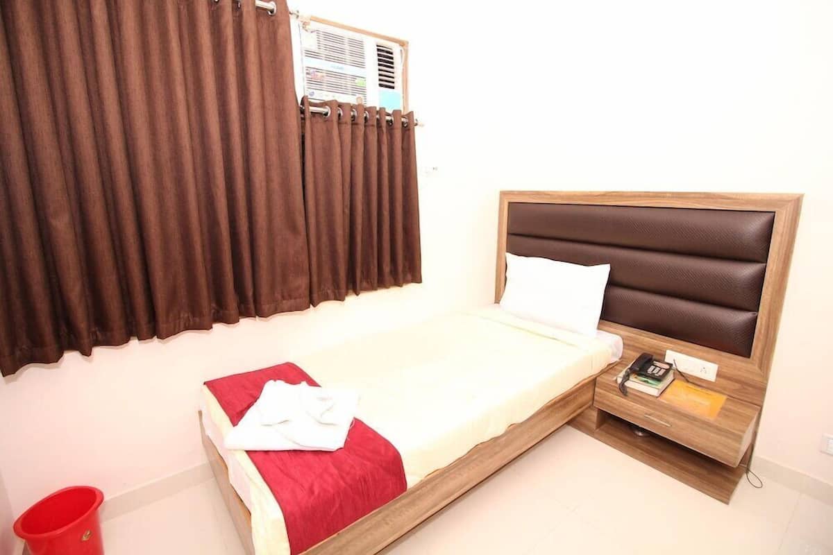 Una cama individual con cabecera de madera, una colcha roja y una almohada blanca. Hay una mesita de noche con un libro, un teléfono y un bloc de notas. Un cubo rojo está en el suelo a la izquierda de la cama.