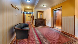 Un pasillo de hotel con alfombra roja estampada, un sillón de cuero negro, un aparador de madera y un ascensor.