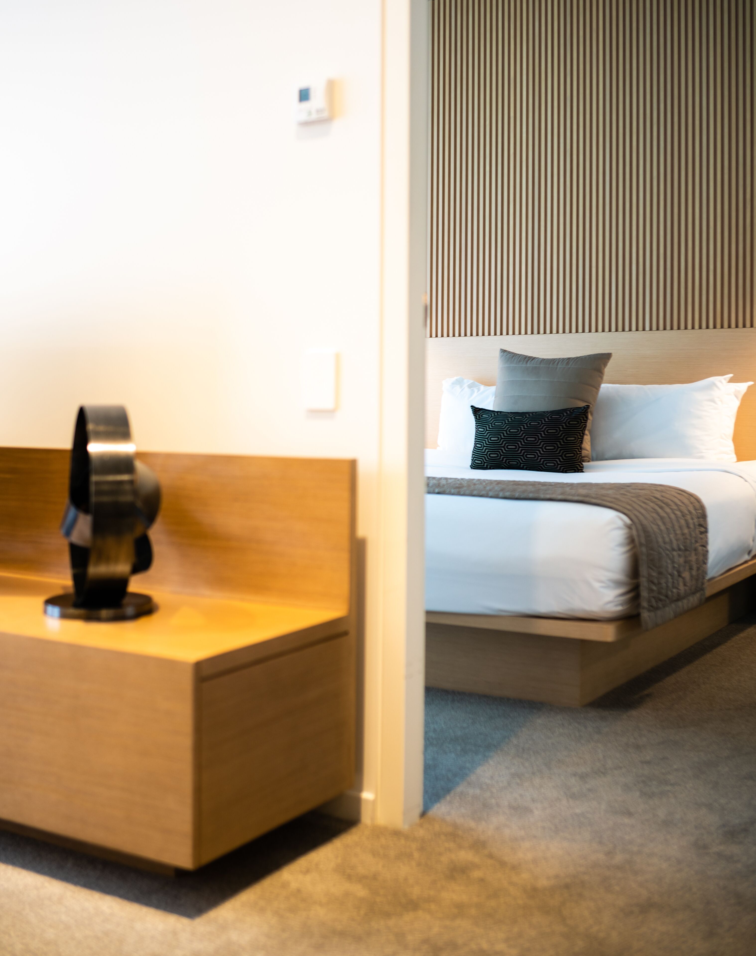 A modern hotel room with a wooden bedside table, a bed with white linens and patterned pillows, and a slatted headboard.