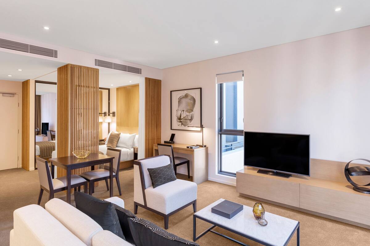 A modern hotel room with a living area featuring a sofa, armchair, coffee table, TV, and a large window.