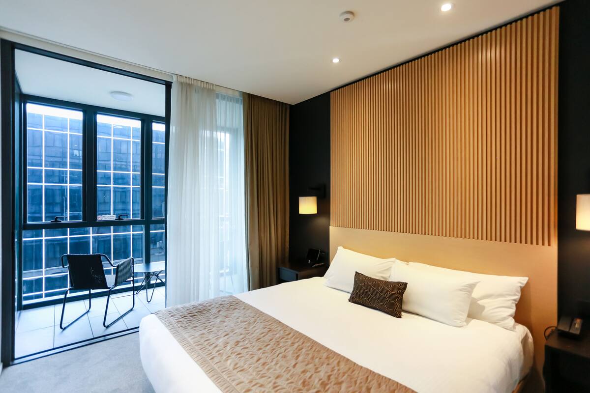 A modern hotel room with a large bed, a view through floor-to-ceiling windows, a balcony with a chair, and a wooden panel wall.