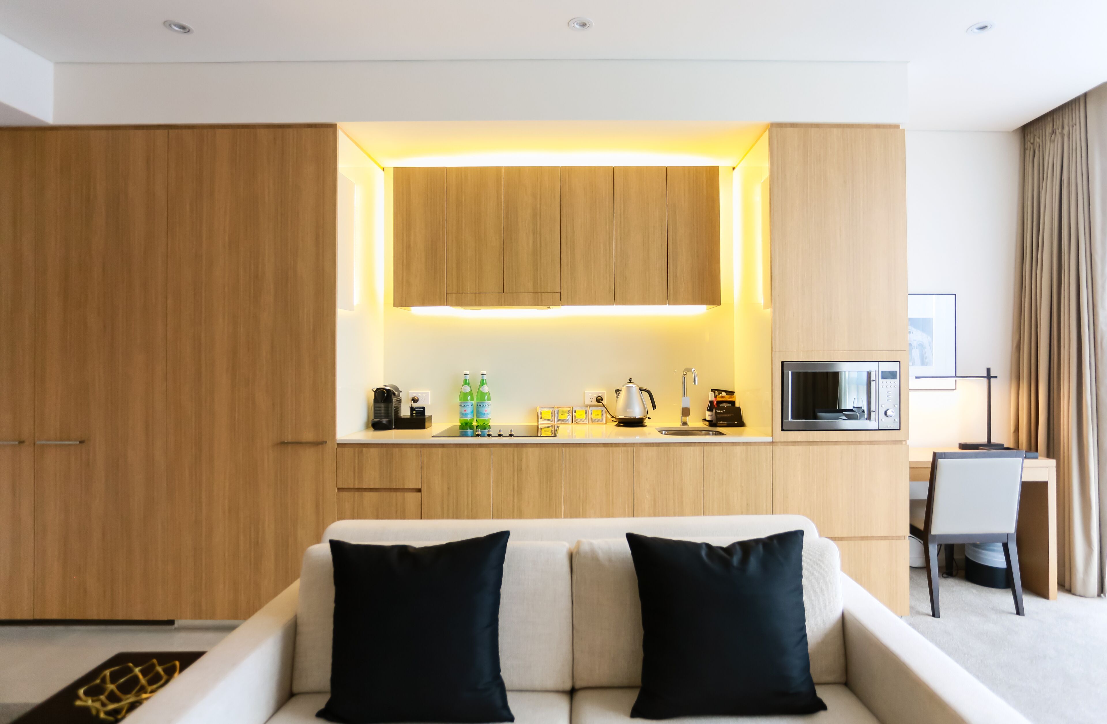 A modern hotel room with a kitchenette, a sofa with black cushions, and a desk with a chair.