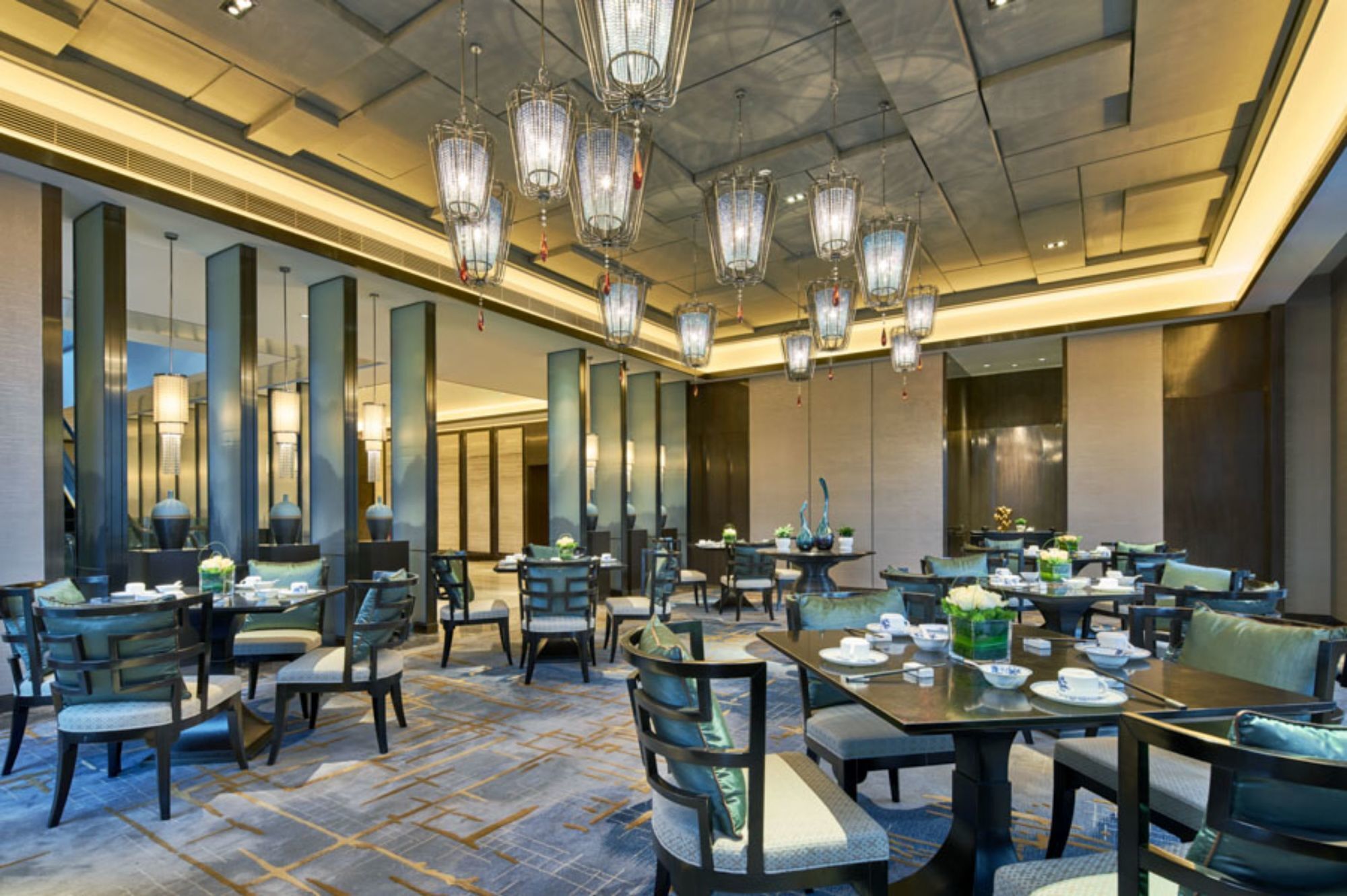 A spacious hotel dining area with multiple tables and chairs, large mirrors, and modern chandeliers.