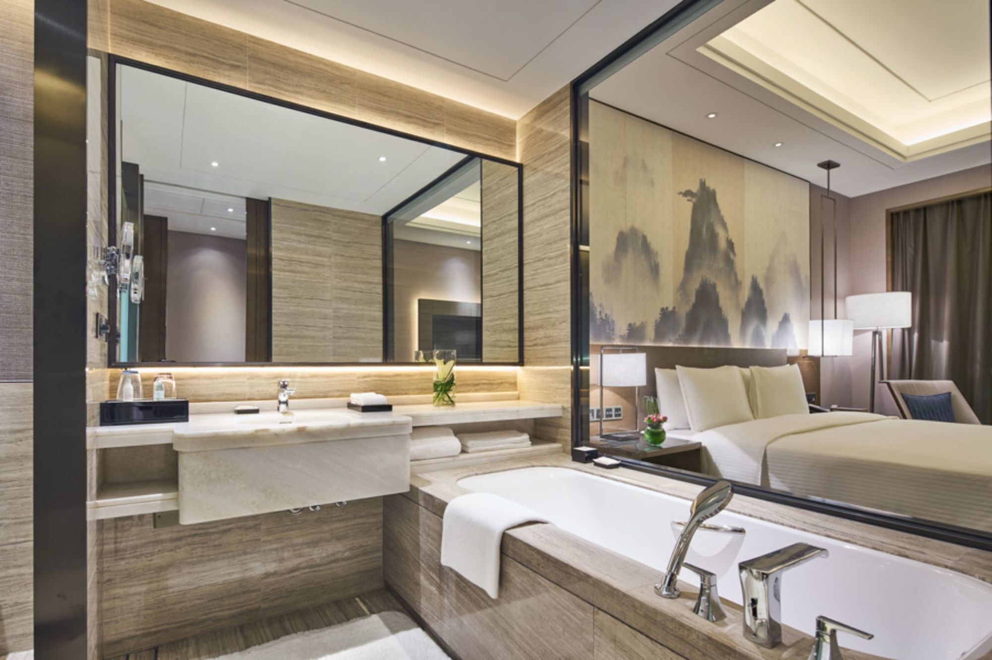 A modern hotel bathroom with a large mirror, a bathtub, a vanity with a sink, and a bed with a headboard.