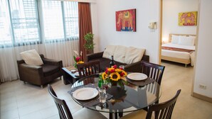 A modern living room with a glass dining table, brown sofas, and a large window with blinds. - Tarntawan Place Apartments (Bangkok Noi)