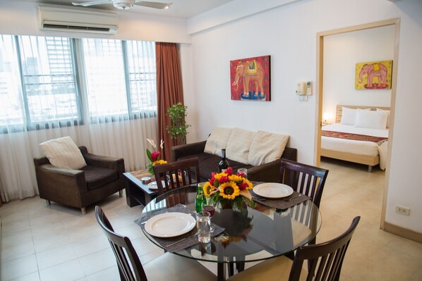 A modern living room with a glass dining table, brown sofas, and a large window with blinds. - Tarntawan Place Apartments (Bangkok Noi)