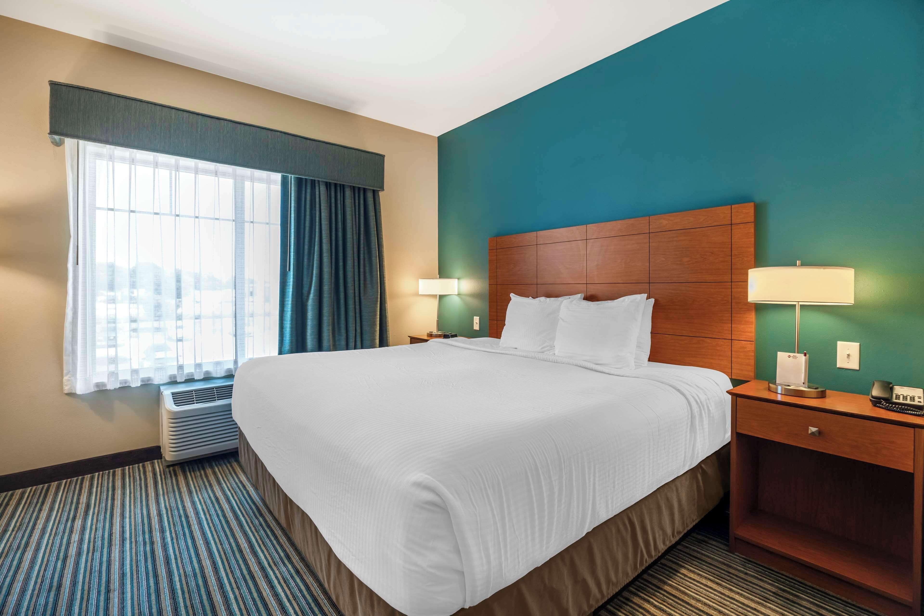 A hotel room with a large bed, a nightstand with a lamp, a window with curtains, and a wall heater.