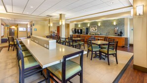 Un comedor de hotel con mesas comunitarias largas y sillas, una estación de café y una cocina al fondo.