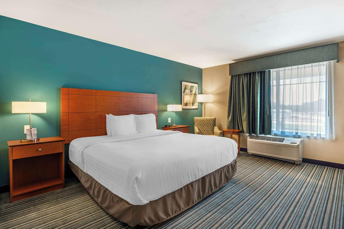 Ein Hotelzimmer mit einem großen Bett, zwei Nachttischen, einem Sessel, einem Fenster mit Vorhängen und einer Klimaanlage.