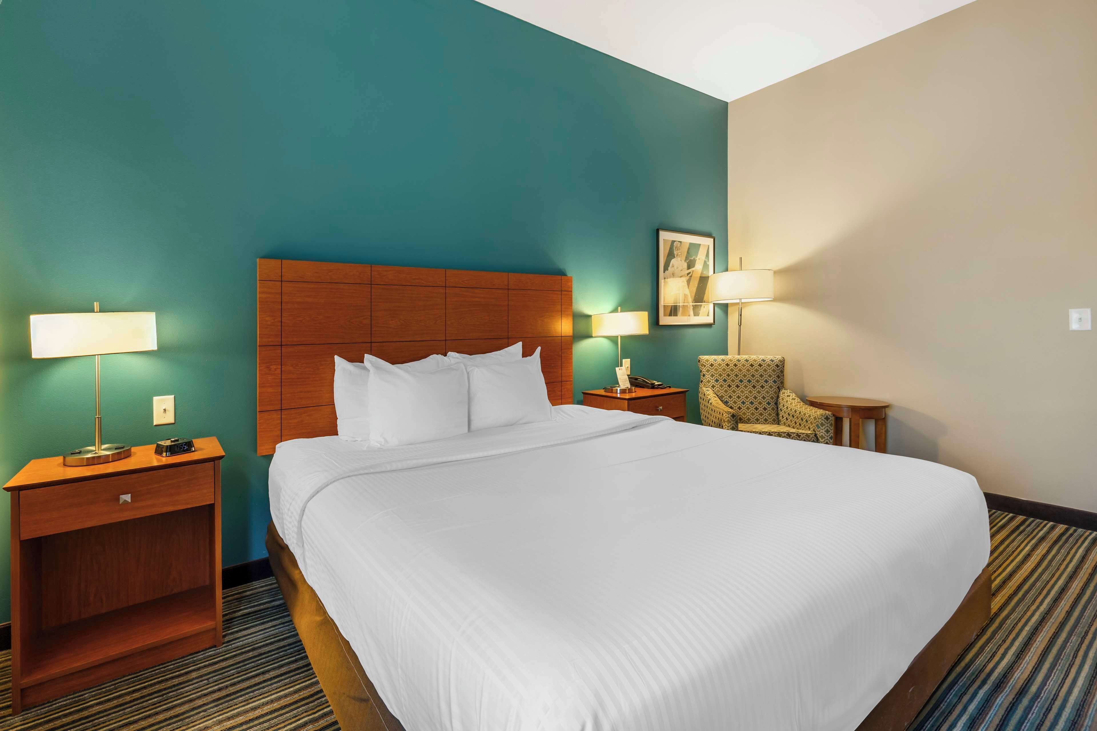 A hotel room with a large bed, a wooden headboard, a nightstand with a lamp, a small table with a lamp, and a patterned armchair.