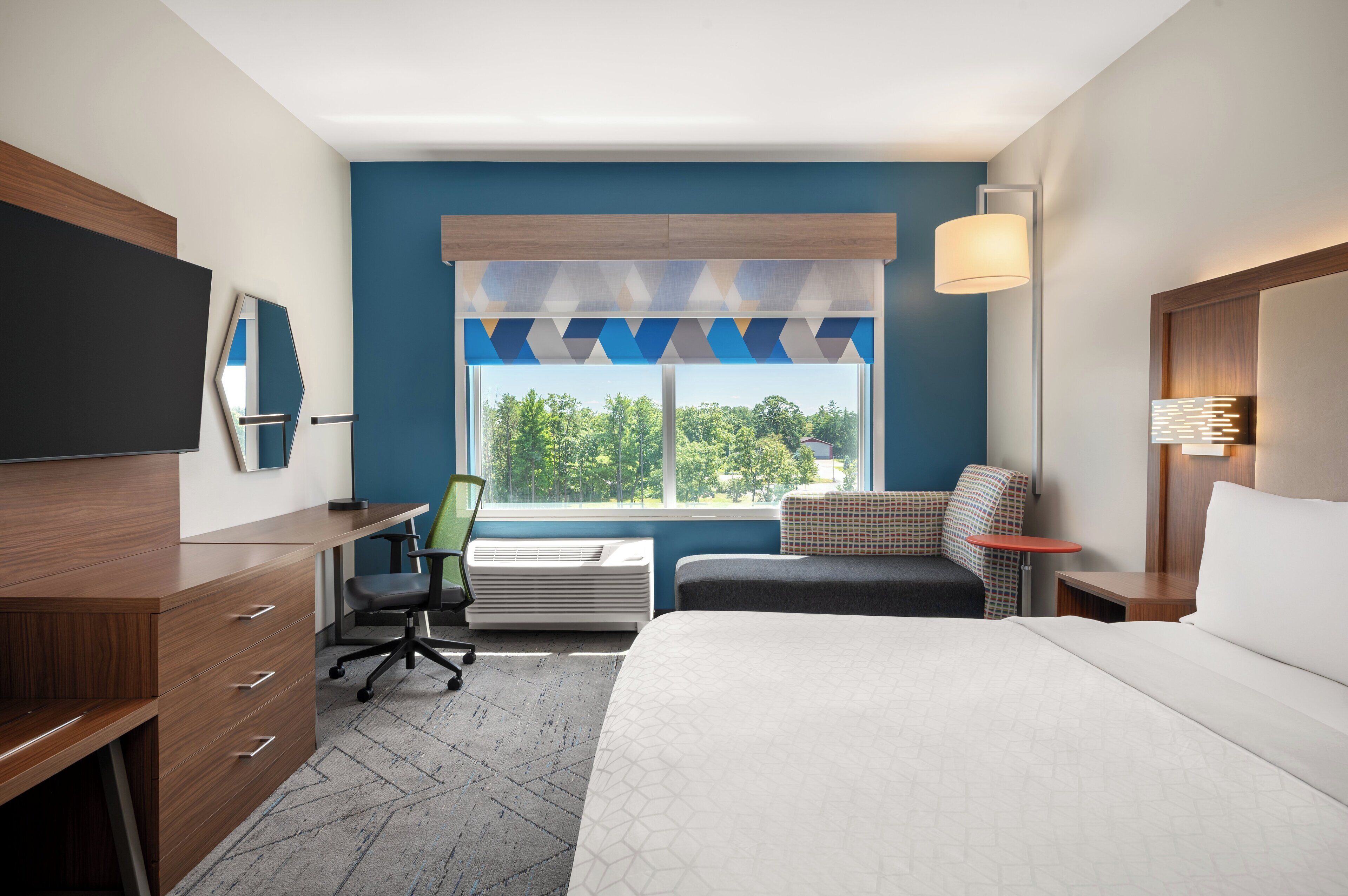 A hotel room with a large bed, a desk with drawers, a chair, a small table, and a window with a view of trees.