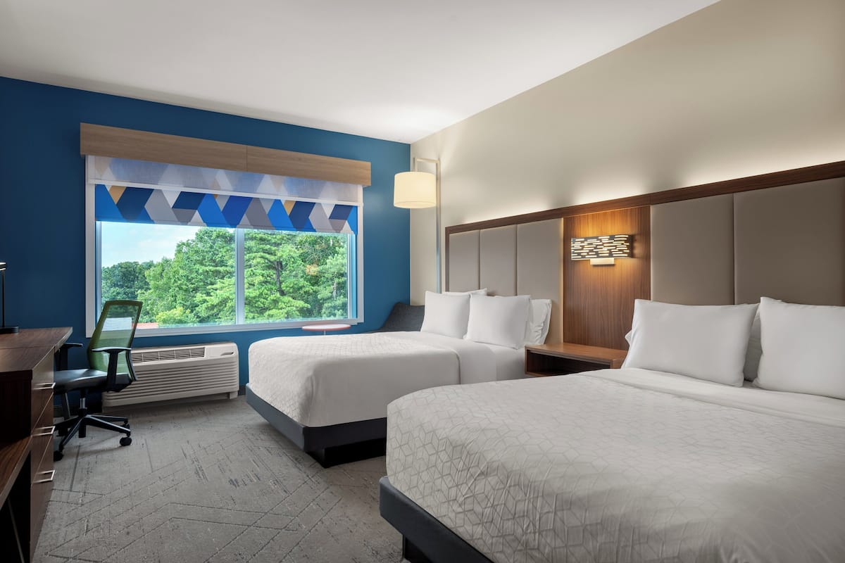 Une chambre d’hôtel avec deux lits, un bureau, une chaise et une grande fenêtre donnant sur un paysage verdoyant.