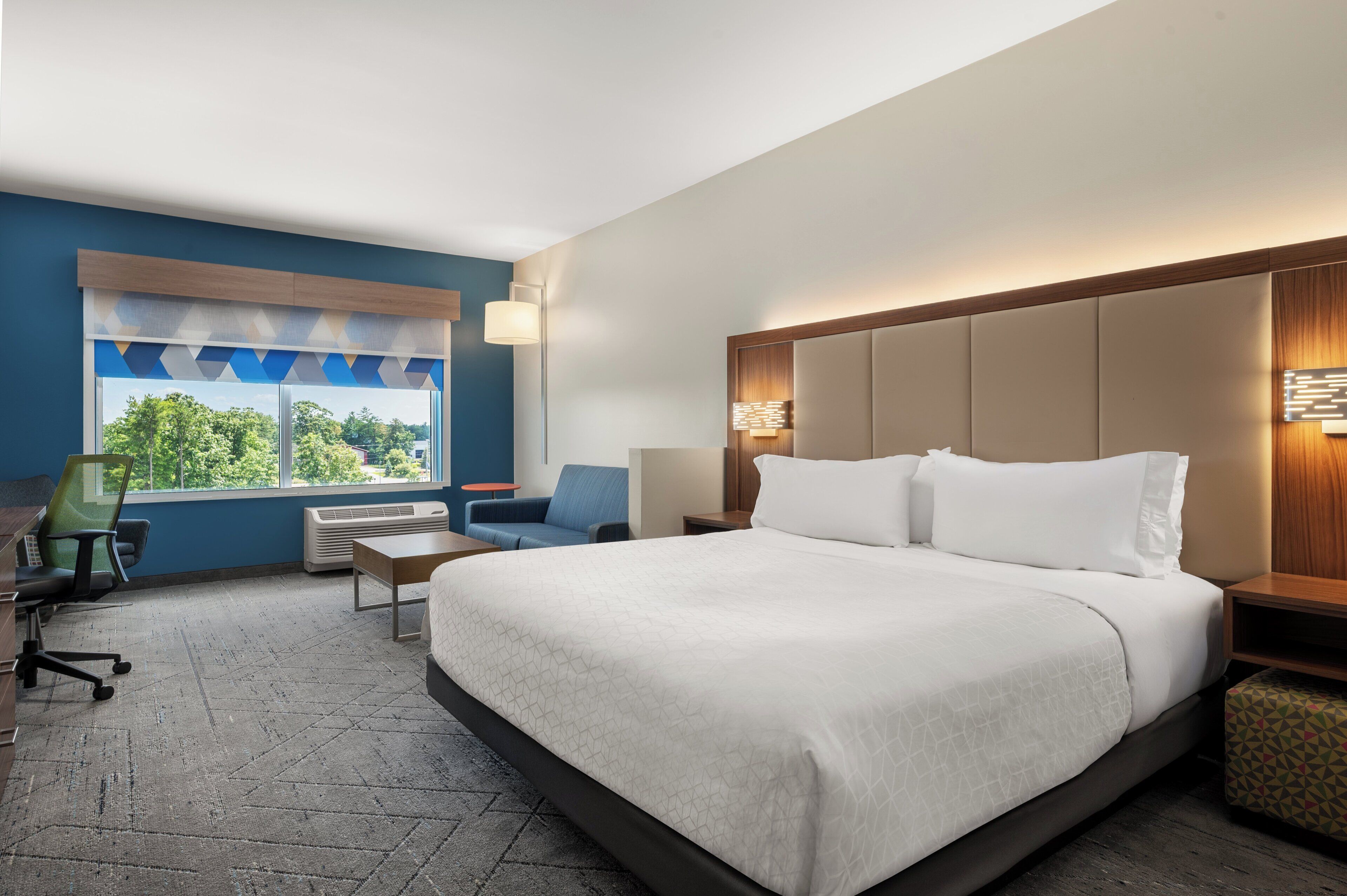 A hotel room with a large bed, a desk, a chair, and a window with a view of trees.
