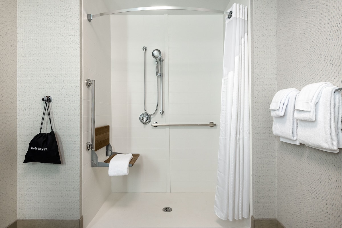 Une douche avec une porte en verre, un porte-serviettes avec des serviettes blanches, et un sac noir sur lequel est écrit « SÈCHE-CHEVEUX », accroché au mur.